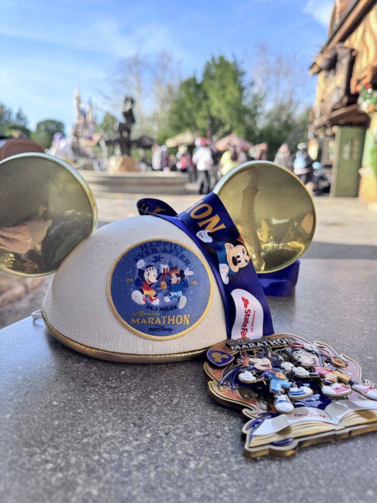 Pale gold "Mickey Mouse ears" hat on a table along with a marathon medal shaped like an open book--both from the 2026 WDW Marathon.